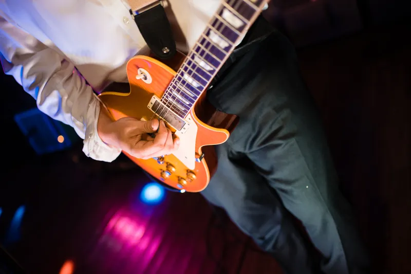 A musician in a white dress shirt plays an orange electric guitar under colorful stage lights during the reception.