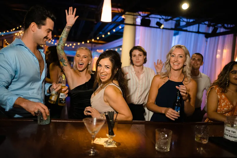 Guests gather at the bar laughing and holding drinks while string lights drape overhead at the indoor venue.