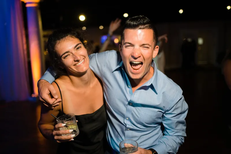 Laurel in a black dress and a guest in a light blue shirt laugh together with drinks in hand under warm overhead lighting.