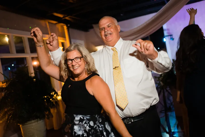 A woman in a black sleeveless dress and a man in a white shirt and yellow tie dance with raised arms during the reception.