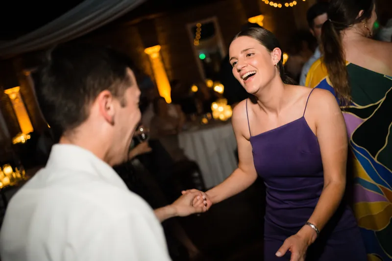 Laurel in a purple tank top laughs while dancing with a guest under warm overhead lighting.