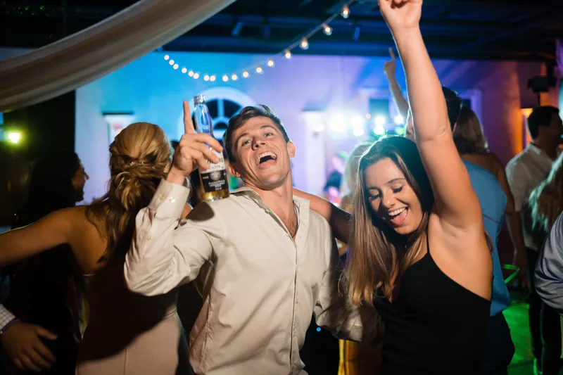 Jake and Laurel dance energetically with friends as guests celebrate around them under blue and green lights.