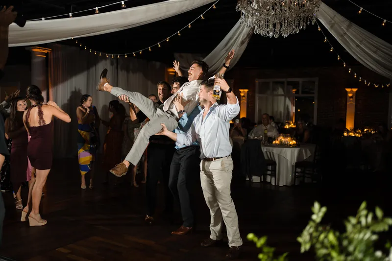 Jake lifts Laurel in the air on the dance floor while guests watch and cheer beneath draped fabric and string lights.