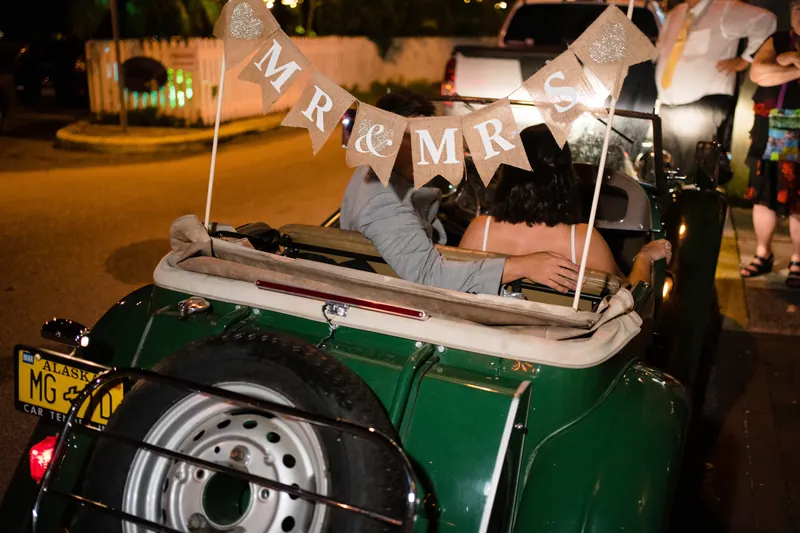 A vintage green convertible displays a "Just Married" banner and packed suitcases for their departure.