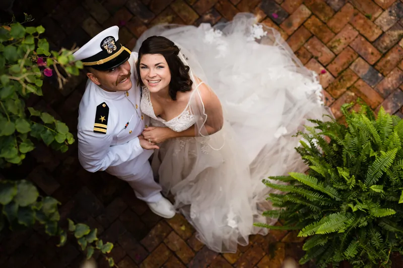 Jake in a white naval uniform and Laurel in her wedding dress pose on a brick courtyard, her veil flowing.