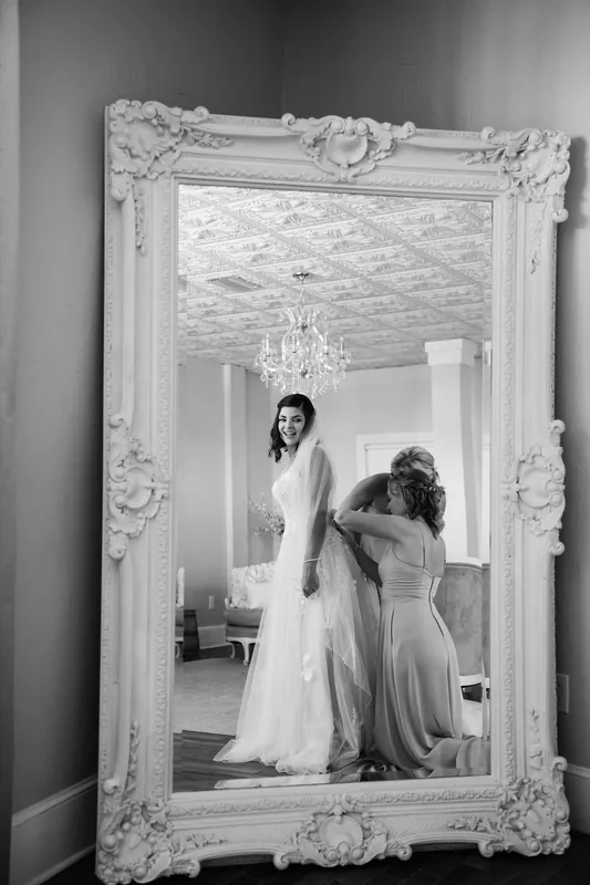 Laurel stands in an ornate white-framed mirror while someone arranges her dress train.