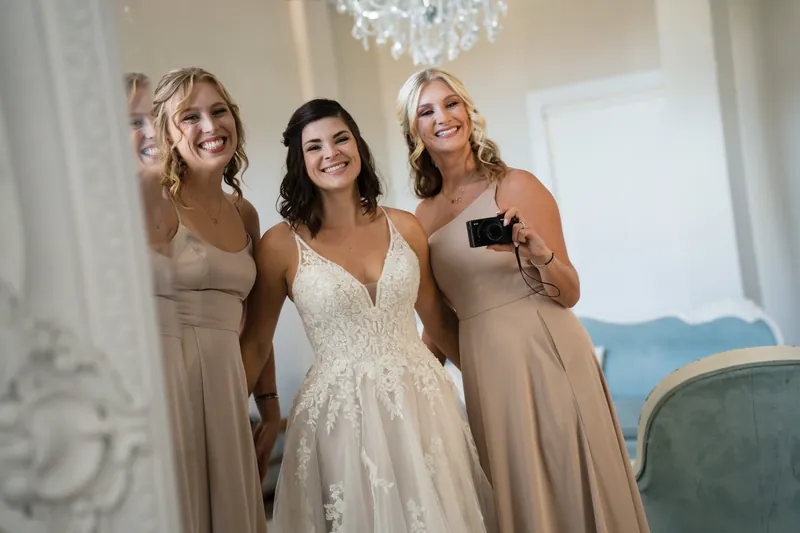 Laurel stands between two attendants in neutral-toned dresses, all smiling in the bridal suite.