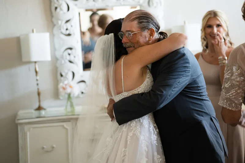 Jake embraces Laurel in her lace wedding dress as guests watch from behind them indoors.