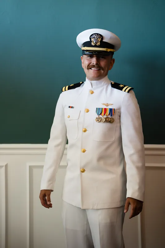Jake stands in dress whites with military medals and insignia pinned to his chest in front of a teal backdrop.