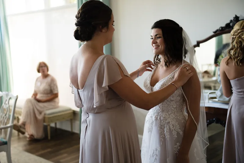A bridesmaid adjusts the back of Laurel's wedding dress while other attendants look on in the bridal suite.