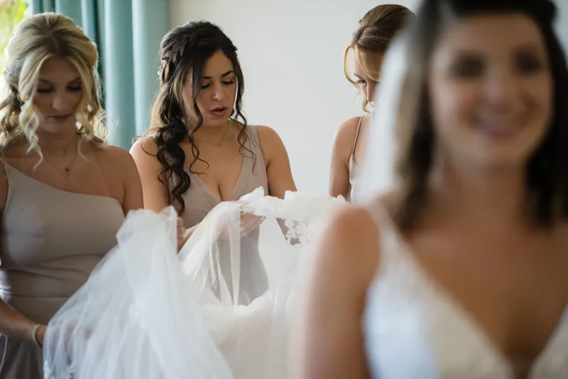 Laurel holds her beaded gown while surrounded by women in neutral-toned dresses examining the intricate beadwork.