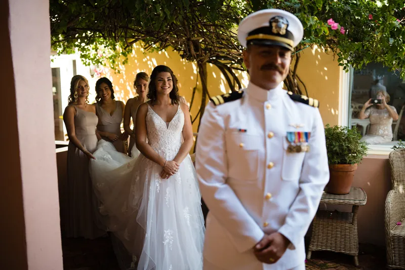 Jake in naval dress whites walks down an outdoor garden corridor as Laurel and bridesmaids follow behind.
