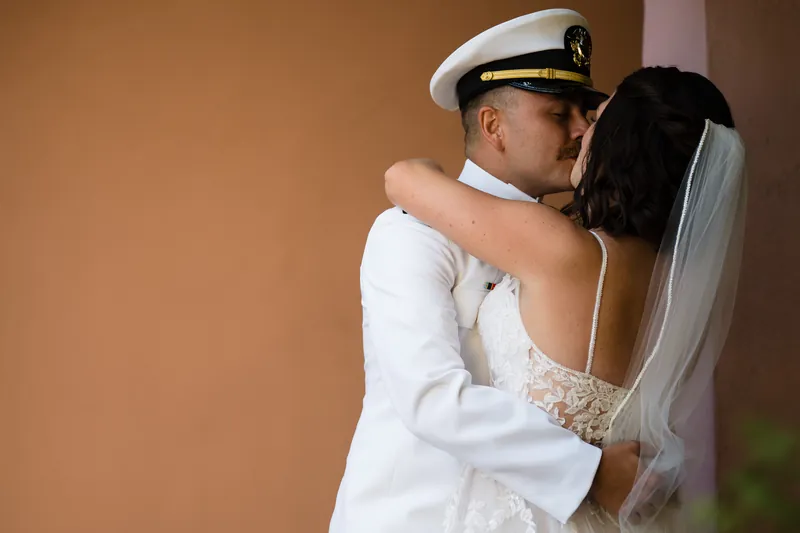 Jake embraces Laurel from behind as she gazes over her shoulder, her lace veil visible against the warm interior light.
