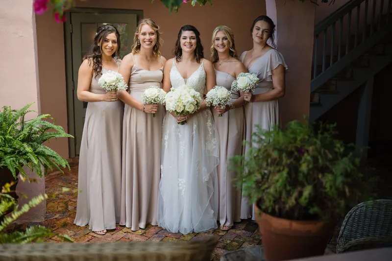 Five bridesmaids in neutral gowns hold white rose bouquets in the courtyard at The White Room, St. Augustine.