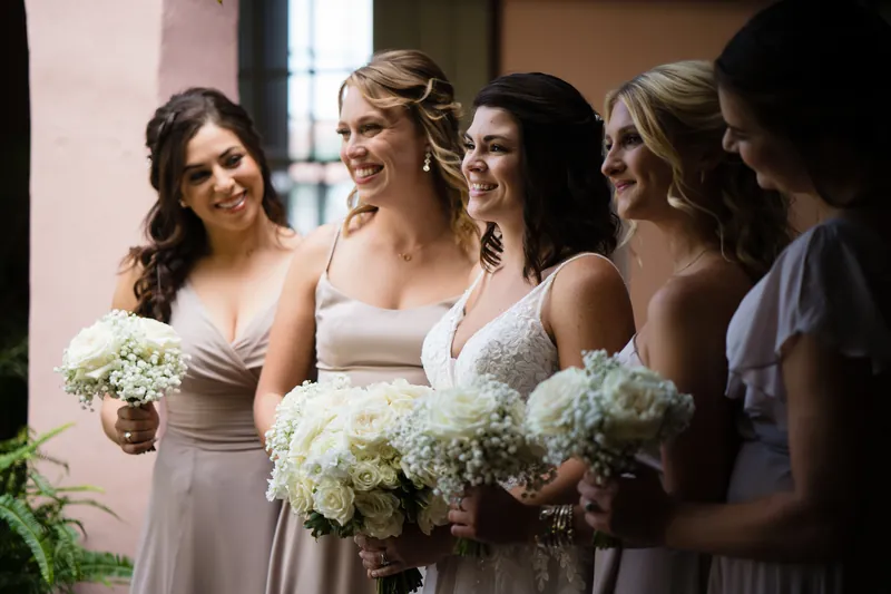 Laurel in a beaded white dress stands with bridesmaids holding white and baby's breath bouquets outside.
