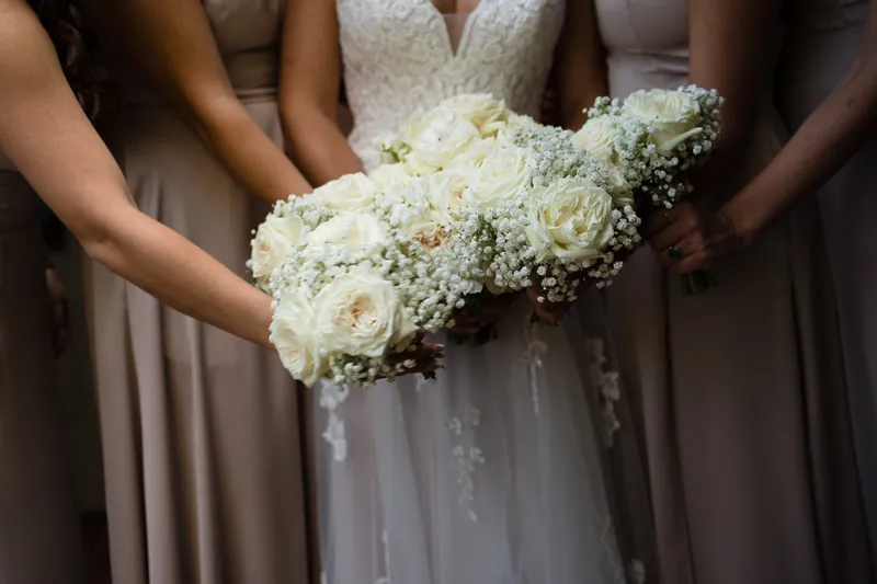 Hands of bridesmaids frame Laurel's white rose and baby's breath bouquet against her detailed gown.