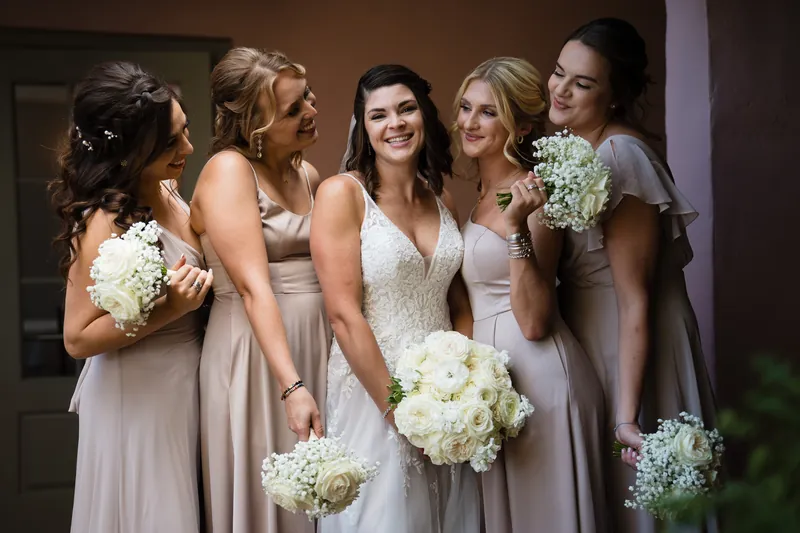 Laurel and four bridesmaids in soft neutral dresses smile together, each holding white floral arrangements.