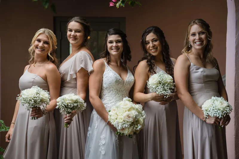 Five bridesmaids in champagne and taupe gowns hold matching white rose bouquets in a courtyard setting.