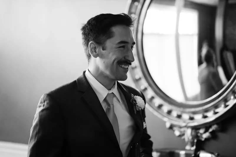 Jake smiles in profile while getting ready at The White Room, wearing a dark suit and boutonniere.