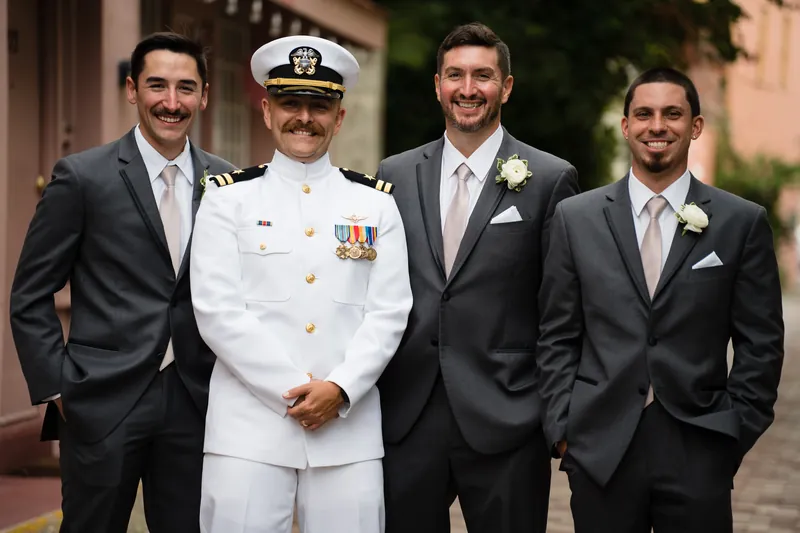 Four men pose together at The White Room, including Jake in a gray suit and a groomsman in naval dress whites.