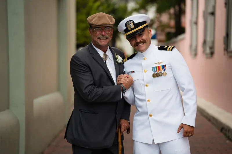 A man in a gray suit shakes hands with a naval officer in white dress uniform outside The White Room.
