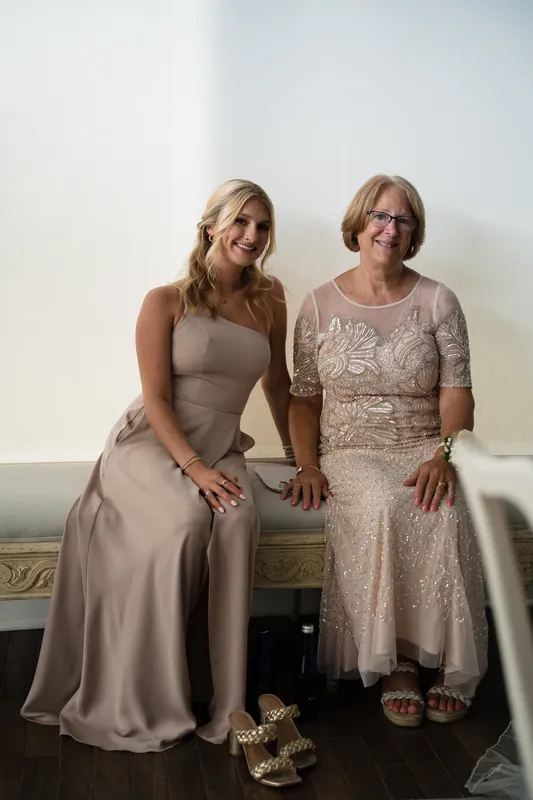 Two women sit together on a bench, one in a taupe gown and the other in a beaded blush dress, smiling at the camera.