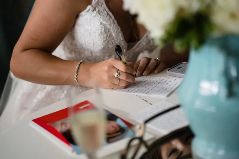 Hands hold a pen over wedding paperwork, the wearer's beaded white dress and bracelet visible as they sign documents.