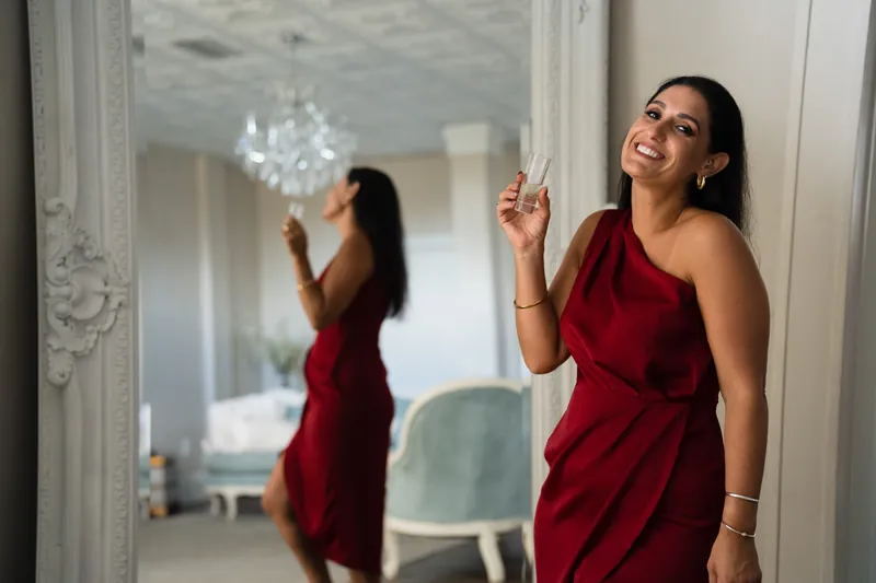 A woman in a burgundy sleeveless dress stands before a mirror, adjusting her hair with a confident expression.