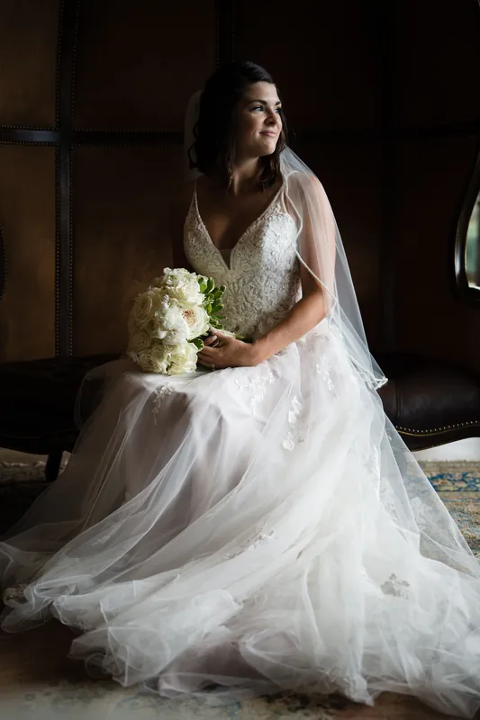 Laurel sits alone in her lace and tulle wedding gown, holding a white bouquet and gazing contemplatively.