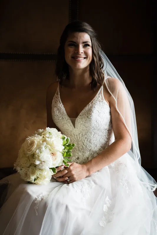 Laurel holds a white rose bouquet and smiles at The White Room, wearing a beaded V-neck wedding dress and veil.