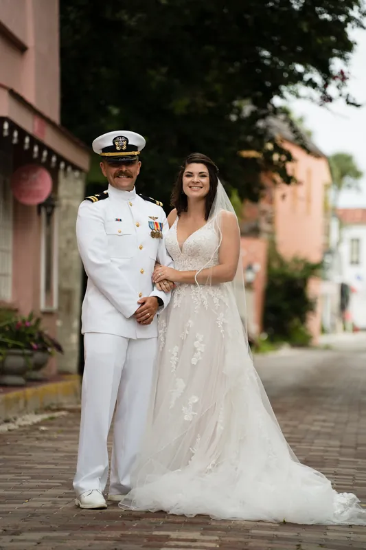 Jake in naval whites and Laurel in her wedding dress stand arm-in-arm on a brick street lined with colonial buildings.