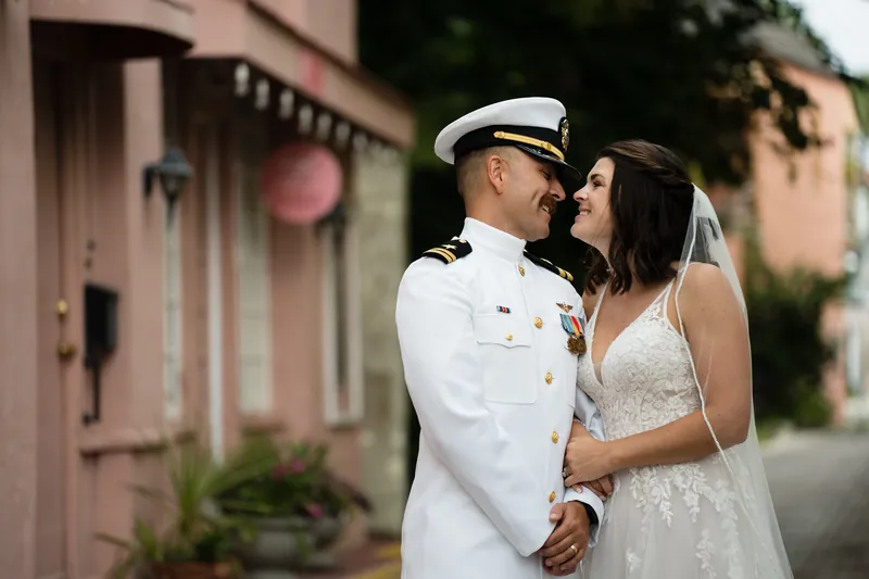 Jake and Laurel embrace outdoors against a pink building, Jake in naval uniform and Laurel in her lace gown.