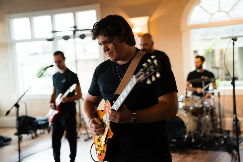 A guitarist in a black shirt performs at The White Room while band members play behind him in the bright, windowed venue.