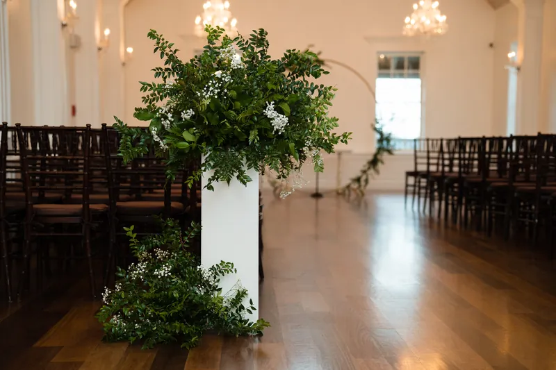 White pedestal with green foliage and white flowers centered in the ceremony aisle between black chairs.