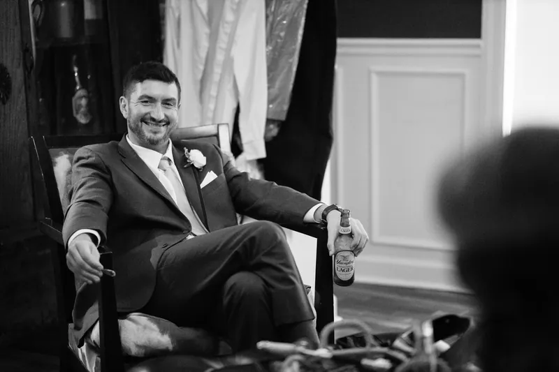 Jake sits relaxed in a dark suit with a white boutonnière, smiling during preparations at the venue.