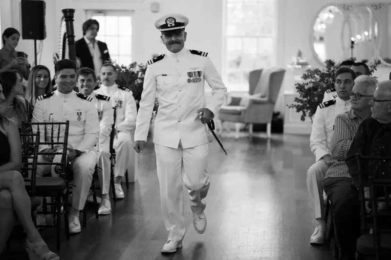 Jake walks down the aisle in white naval dress uniform with medals, flanked by seated guests during the ceremony.