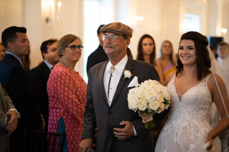 Laurel's father walks her forward while holding her bouquet, with guests visible on either side in the reception space.