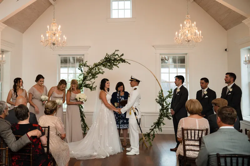 Laurel in a beaded white gown stands with Jake in naval dress at the altar beneath a leafy arch.