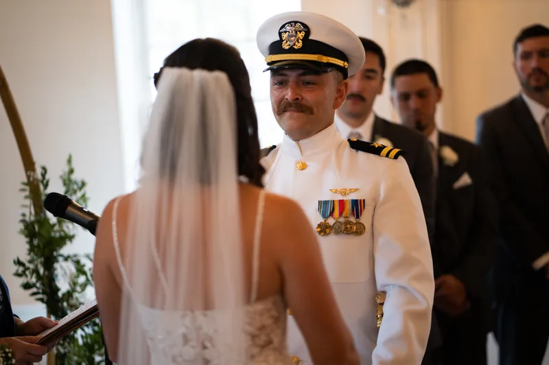 Jake in a white naval uniform with medals faces Laurel, whose veil flows behind her during their ceremony.