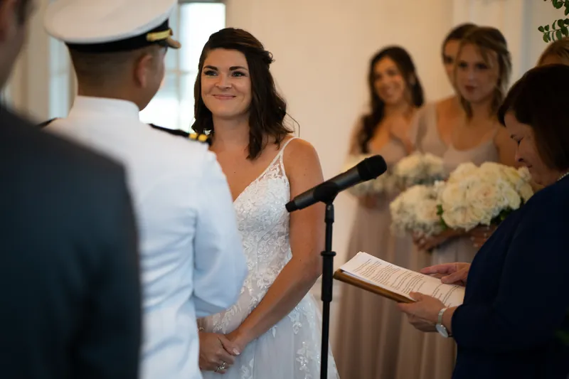 Laurel beams at Jake while speaking at a microphone, surrounded by family members in the elegant reception space.