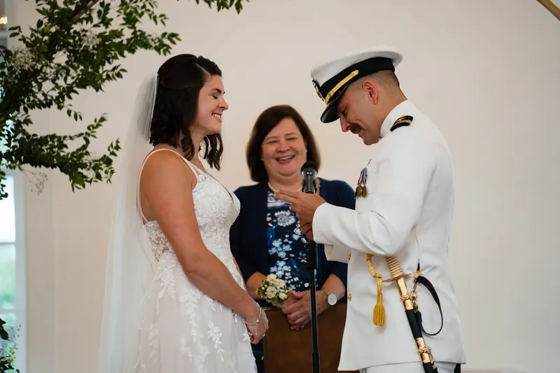 Laurel in a beaded white gown stands between an officiant in black and Jake in naval whites at The White Room.