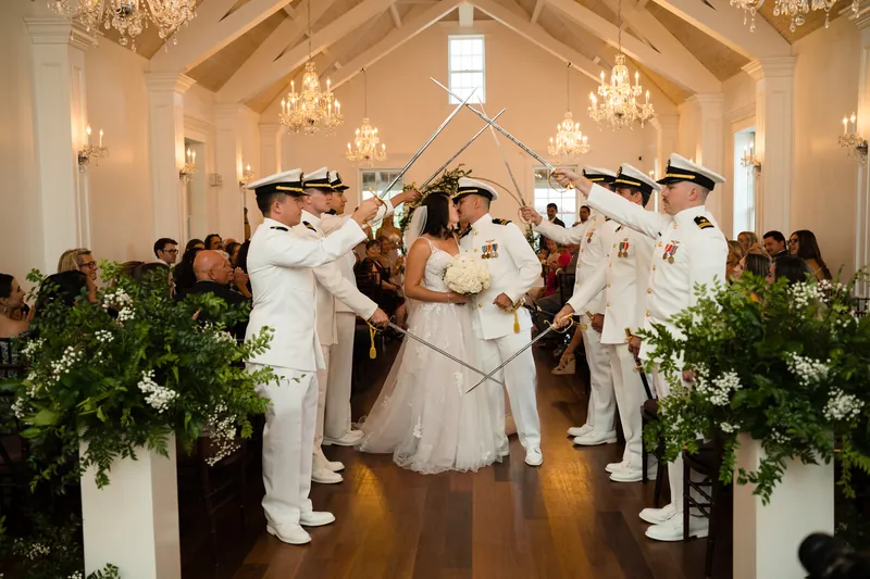 Six naval officers raise crossed swords overhead as Laurel and Jake pass beneath in The White Room's chandelier-lit hall.