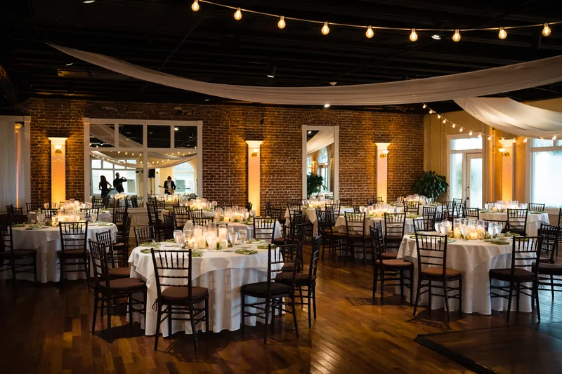 Round tables set with white linens fill The White Room's brick reception space, strung with warm overhead lights.