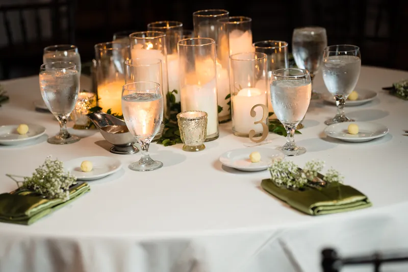 Tiered white cake with floral piping sits on a wooden table surrounded by candlelit glasses and greenery.