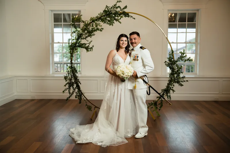 Laurel and Jake stand under a greenery-draped arch in white wedding attire, holding a white bouquet.