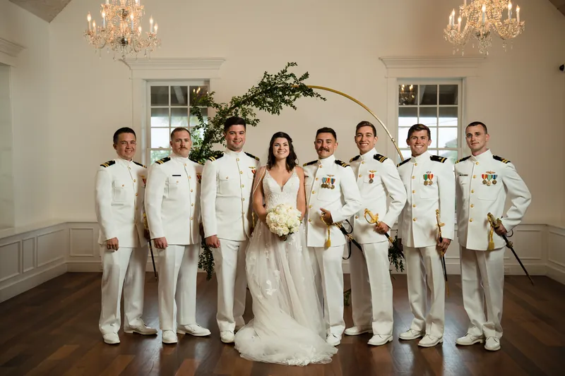 Laurel in a flowing white gown stands with eight groomsmen in cream suits beneath a gold arch.