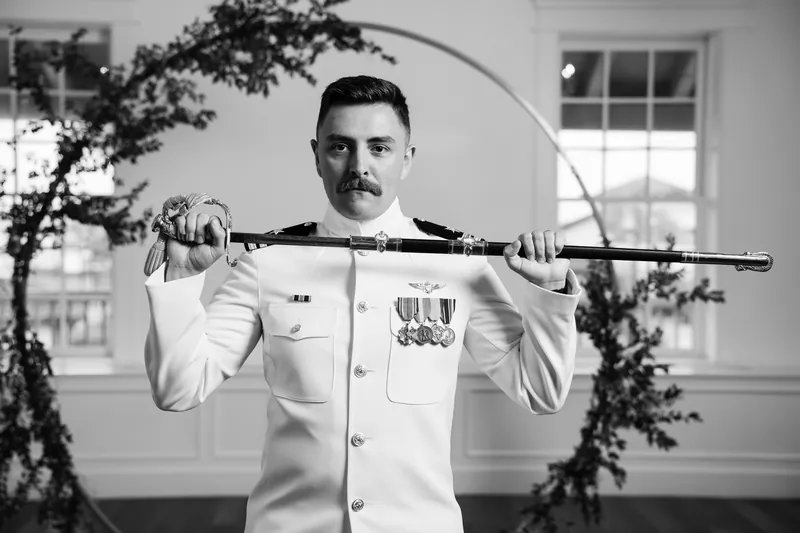 Jake in white naval dress uniform holds a sword across his shoulders, framed by draped greenery and tall windows.