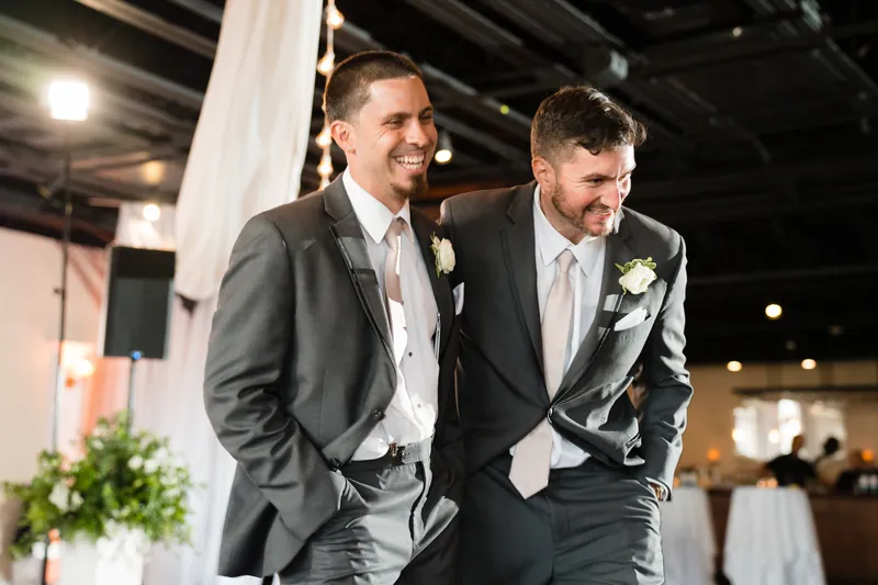 Two men in gray suits with boutonnières walk together, smiling outdoors under industrial ceiling beams.