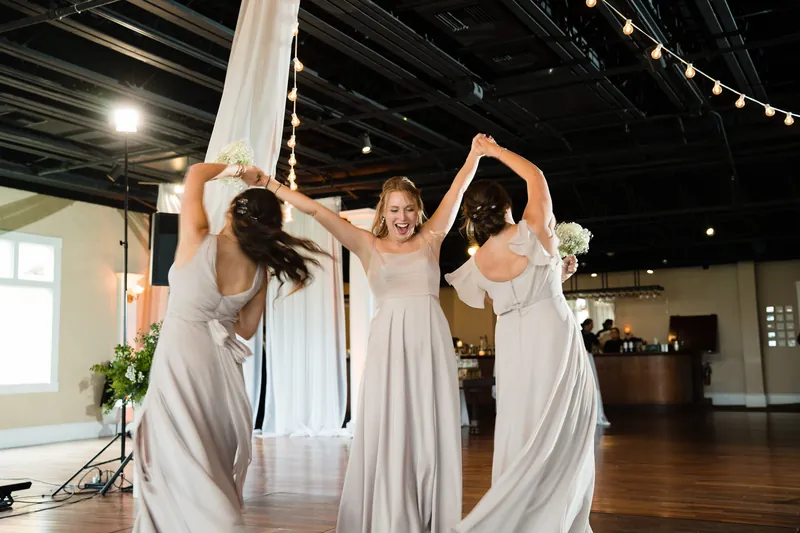 Three women in flowing pale dresses dance with arms raised, holding sheer white fabric between them on the dance floor.