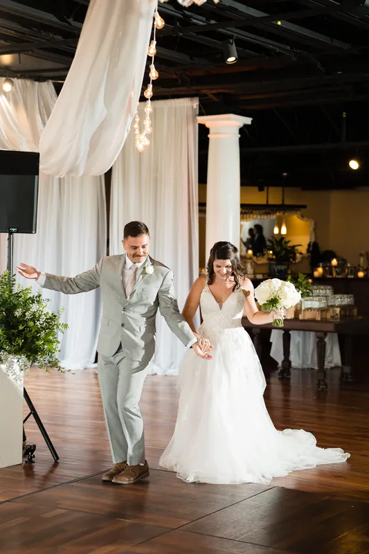 Jake and Laurel walk hand in hand under draped white fabric with greenery and warm lighting.